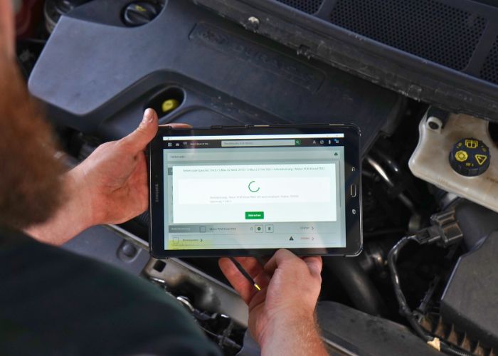 Werkstatt Mitarbeiter führt eine Motordiagnose an einem Fahrzeug aus und hält dabei ein Tablet in der Hand.